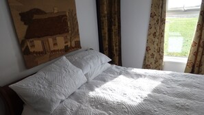 Una cama bien hecha con cubrecama acolchado, un cuadro de una casa en la pared y cortinas con estampado floral.