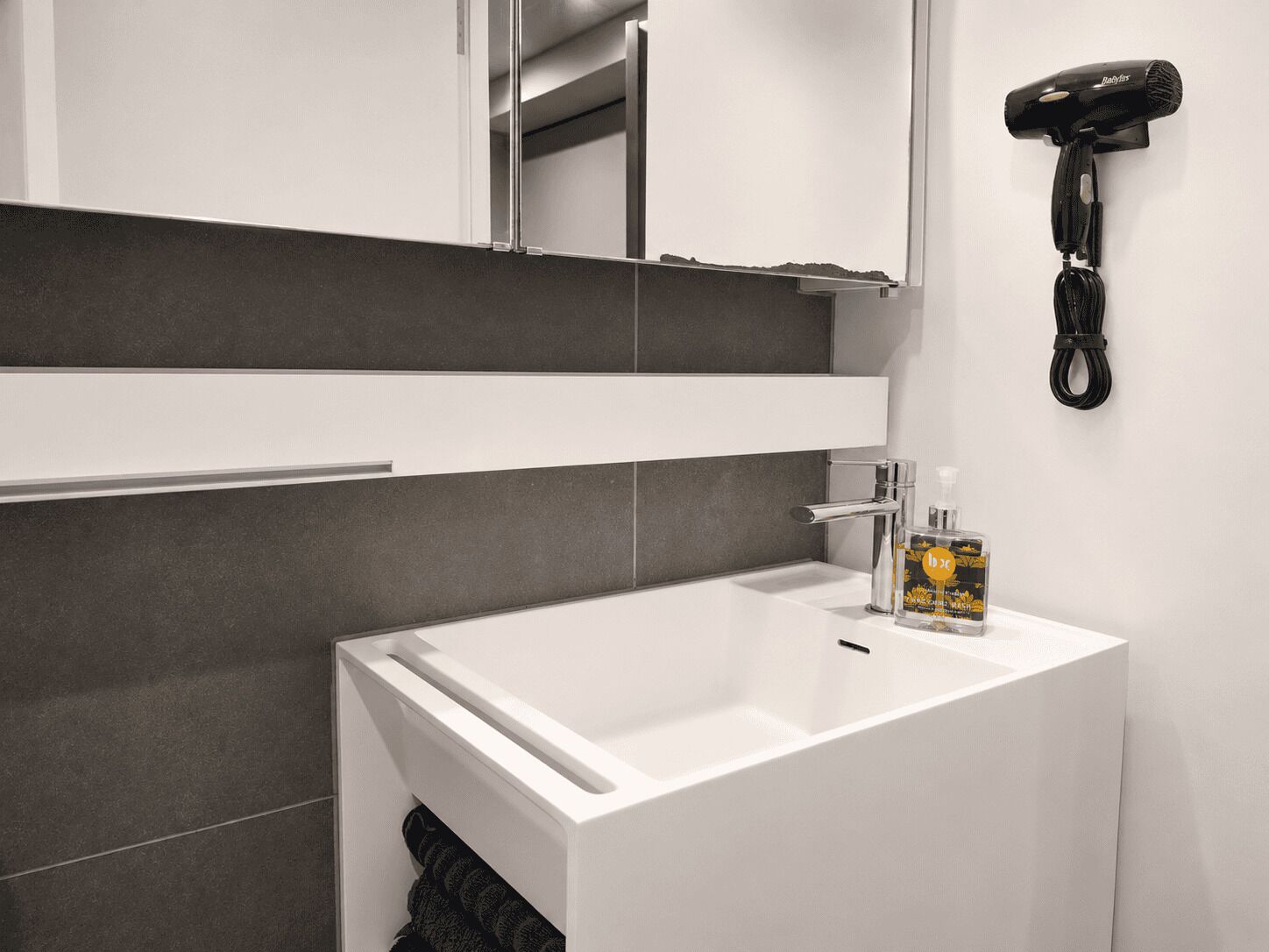 A modern bathroom with a white sink, a mirror, and a hairdryer mounted on the wall.