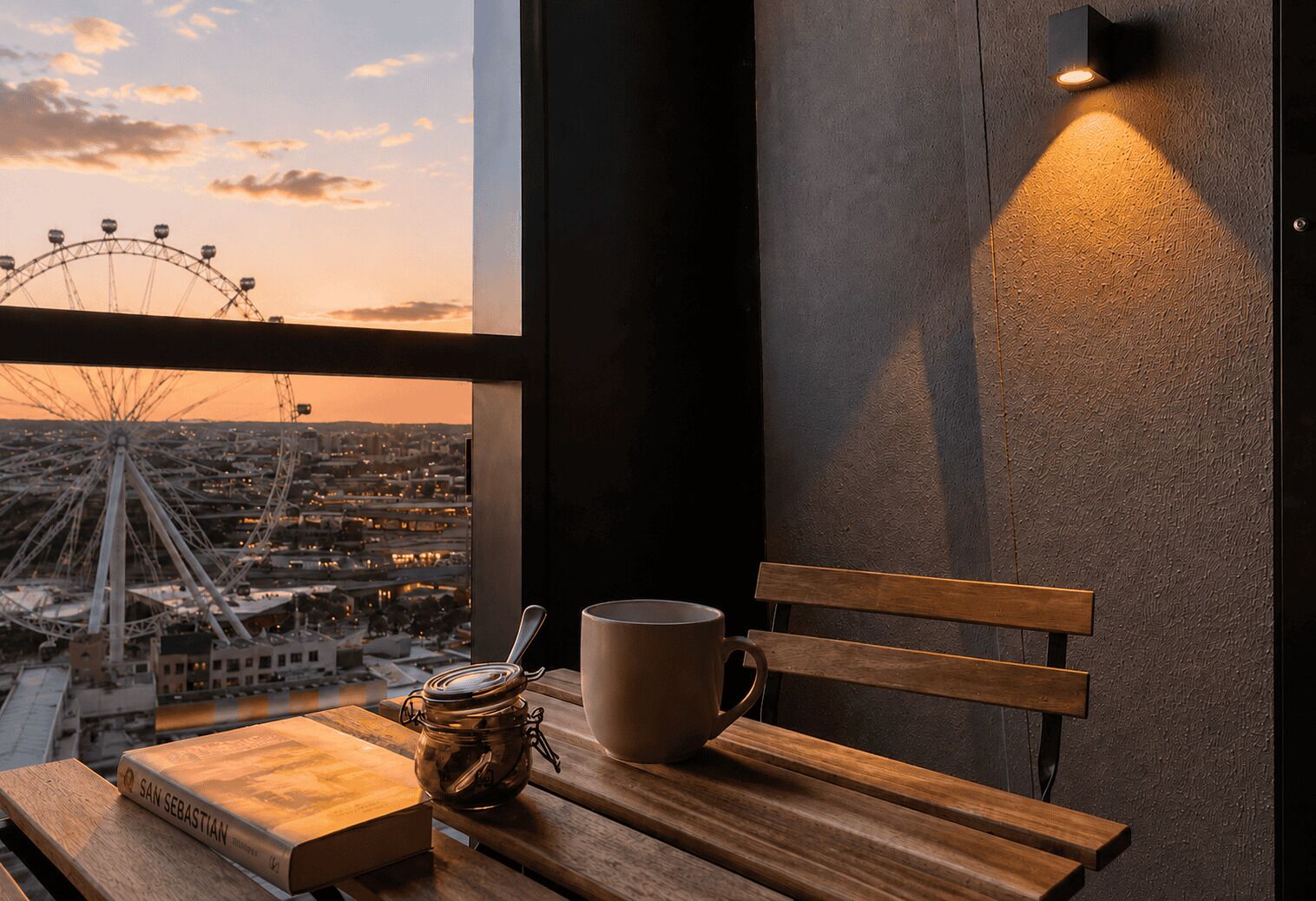 Un banco de madera con una taza, un frasco, un libro con el título 'SAN SEBASTIAN' y vistas a una noria a través de una ventana.