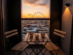 A balcony with a wooden table and two chairs, overlooking a cityscape with a Ferris wheel during sunset. - 23rd-Flr Docklands Views, Pool, Jacuzzi, Free Tram (Docklands)