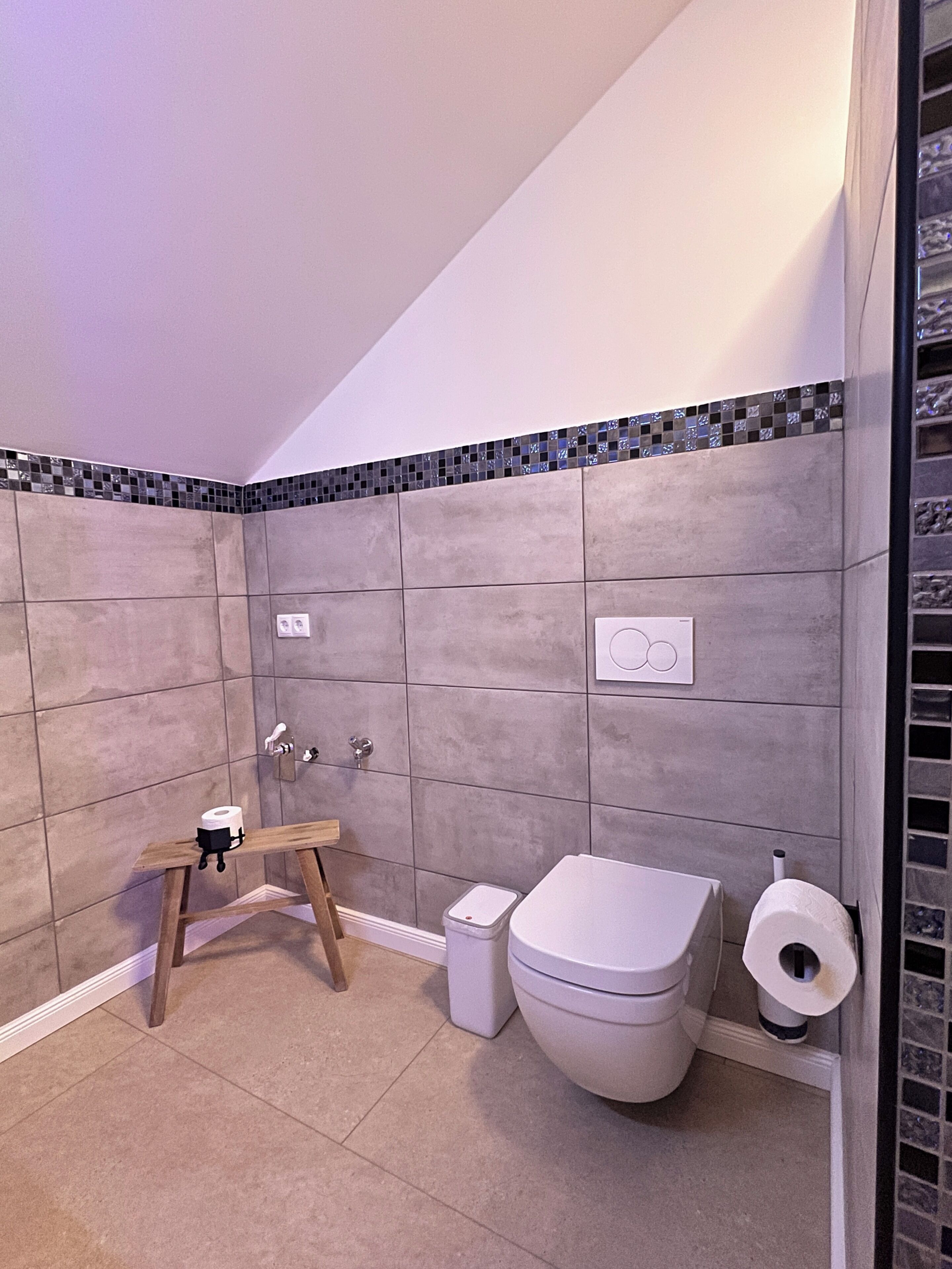 A bathroom with grey tiles, a white toilet, a small wooden stool, a trash bin, and a toilet paper holder.