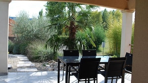 Un patio al aire libre con una mesa y sillas de vidrio, rodeado por un jardín con una palmera y diversas plantas.