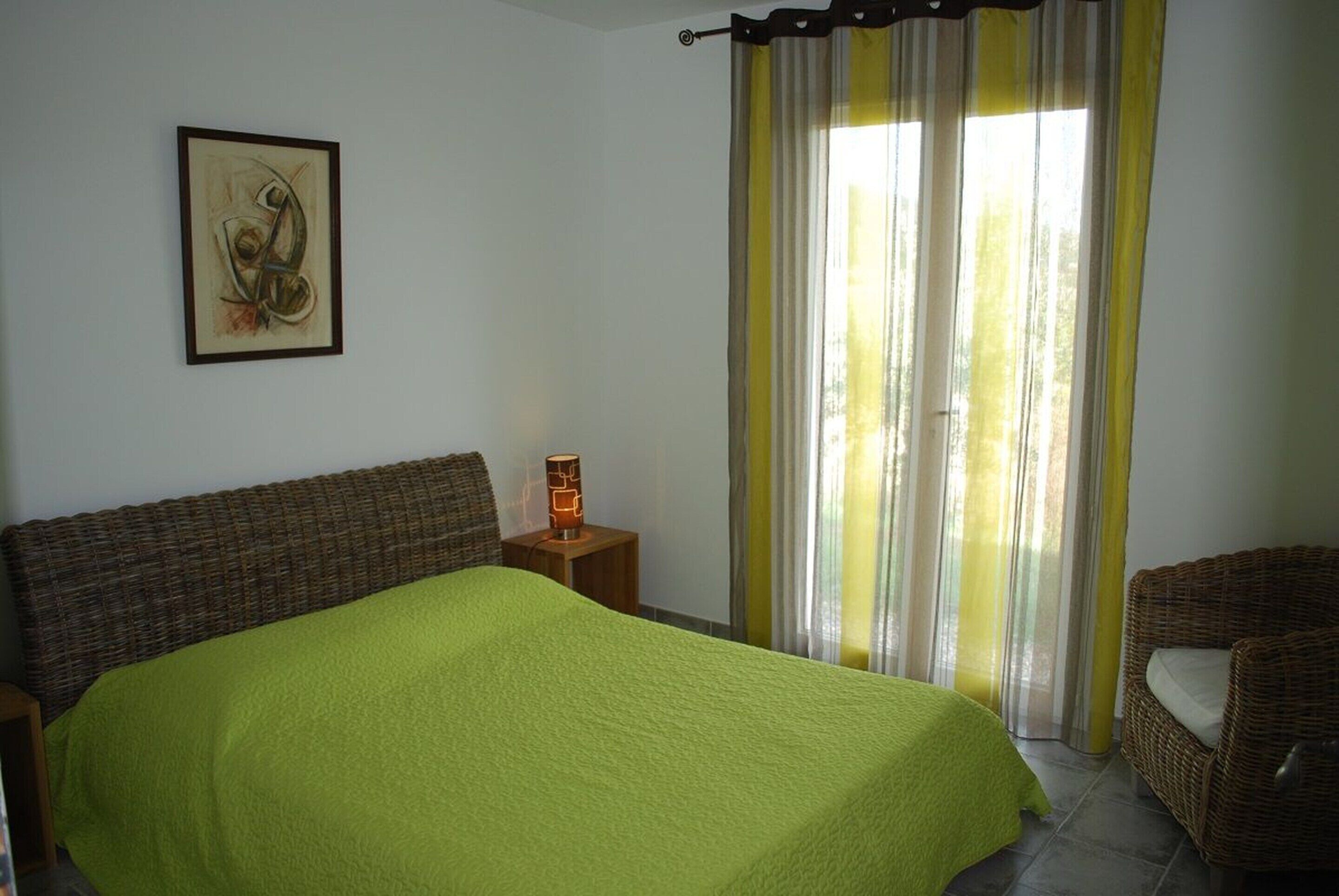 A room with a bed, a wicker chair, a painting on the wall, and a window with curtains.