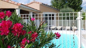 Una piscina con cerca blanca, rodeada de flores rosadas y un edificio con techo de tejas.
