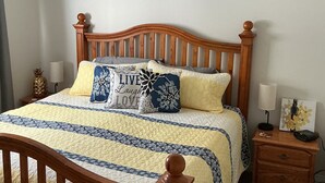 A bed with a wooden headboard, a quilt, and decorative pillows. There is a nightstand with a lamp and a small table with a picture.