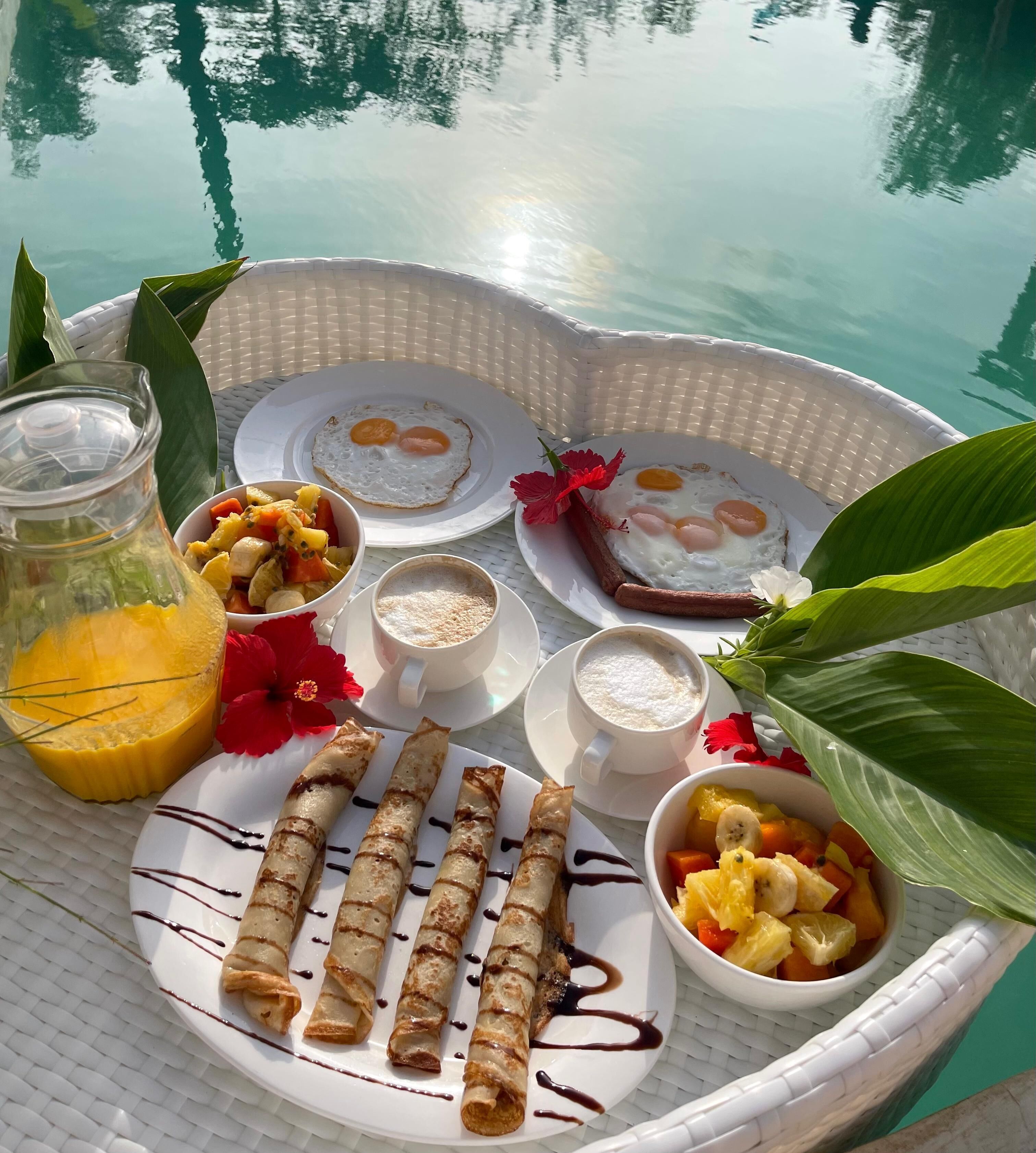 A breakfast tray with eggs, fruit, crepes, and beverages by a pool.