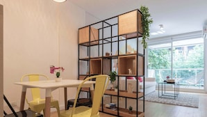 Una sala de estar moderna con una mesa blanca, sillas amarillas, una estantería negra y vistas a zonas verdes a través de amplios ventanales.
