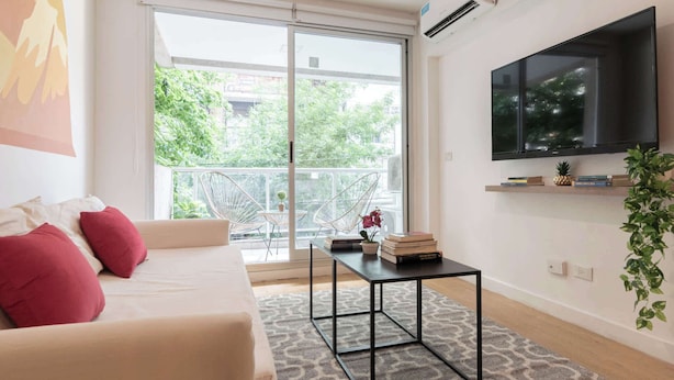 Una sala de estar moderna con sofá, una mesa de centro, un televisor montado en la pared y un balcón con vistas a zonas verdes.