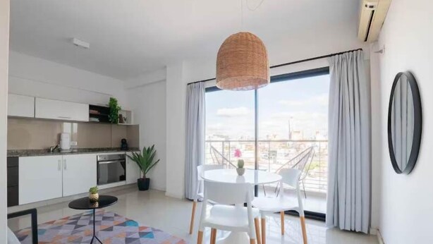 Eine moderne Küche mit weißen Schränken, eine Essecke mit einem runden Tisch und Stühlen sowie ein großes Fenster mit Vorhängen.
