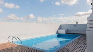 Ein Schwimmbecken auf einer Terrasse mit Holzboden und Metallgeländern, umgeben von einer weißen Mauer und blauem Himmel.