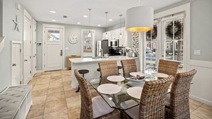 A modern kitchen with a central island, white cabinetry, and a dining area with wicker chairs and a glass table. - NEW to VRBO-FREE Golf & Activities ~Golf Cart~Hot tub, Pool, Gym (Miramar Beach)