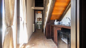 Un pasillo estrecho con una zona de cocina a la derecha, que incluye un fregadero y armarios. Se ve un radiador a la izquierda, y una ventana con cortinas al mismo lado.