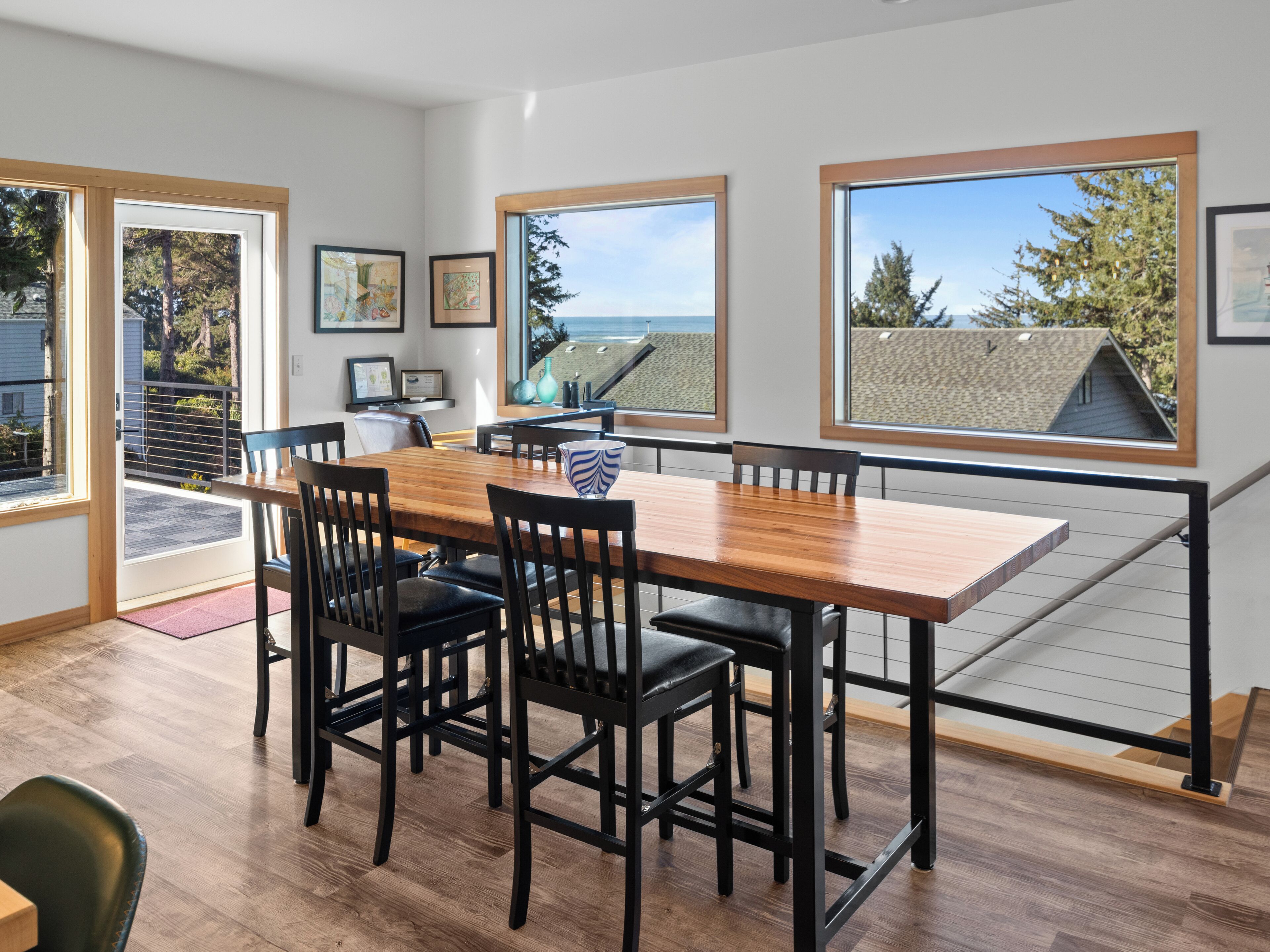 A dining area with a wooden table and black chairs, large windows with a view of the beach, and a balcony with a railing.
