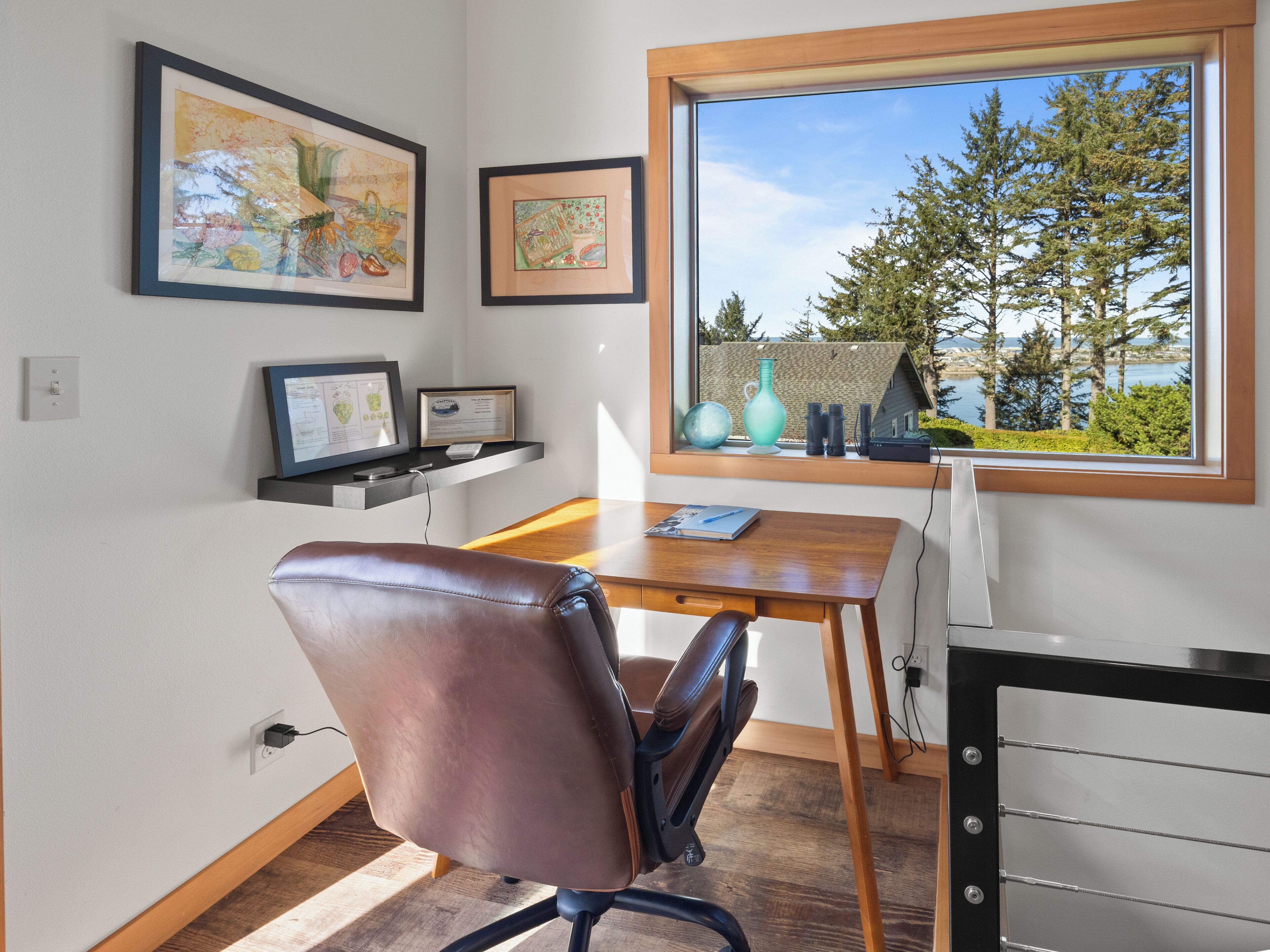 A modern office space with a desk, chair, framed artwork, and a window with a view of trees and a building.