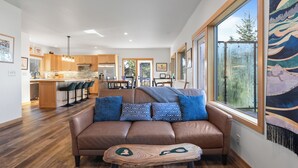 Een moderne woonkamer met een bruine leren bank, een houten salontafel en een groot raam met uitzicht op bomen.