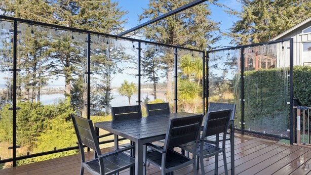 Een terras buiten met een tafel en stoelen, omgeven door glazen reling, met uitzicht op water en bomen.