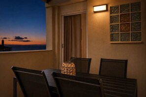 Ένα μπαλκόνι με ένα τραπέζι και καρέκλες, ένα διακοσμητικό καλάθι από ψάθα, μια πόρτα με κουρτίνες και ένα φωτιστικό τοποθετημένο στον τοίχο.