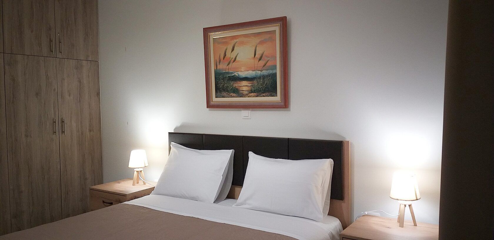 Un dormitorio con una cama, dos lámparas de noche, una mesita de noche, un armario de madera y un cuadro enmarcado en la pared.