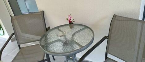 Une table ronde en verre, accompagnée de deux chaises en métal ajouré, d’une petite plante en pot et d’une vue sur un balcon.