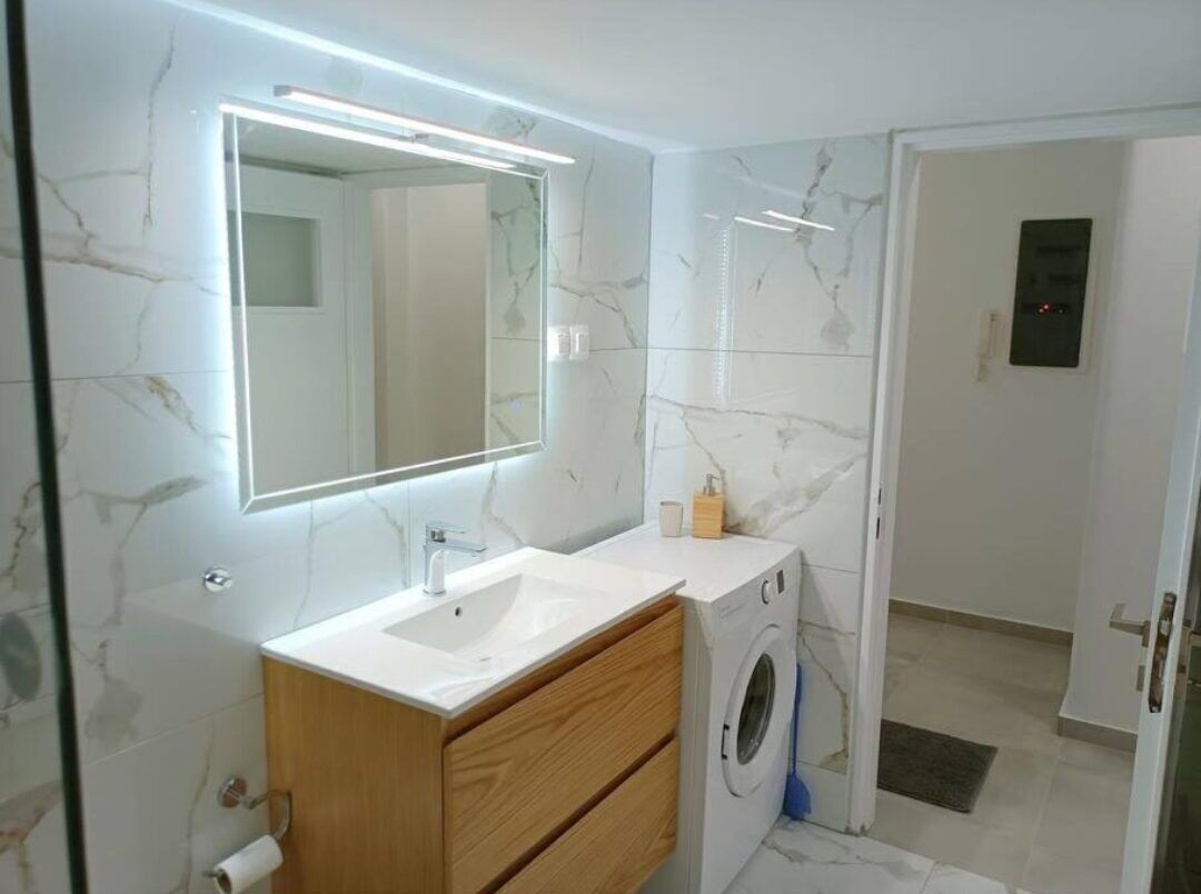 Un baño con pared de mármol, un lavabo, un espejo, una lavadora y una puerta a otra habitación.