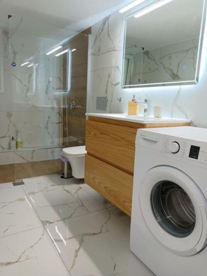 A modern bathroom with a glass shower enclosure, a wooden vanity, a toilet, and a front-loading washing machine.
