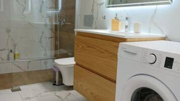 A modern bathroom with a glass shower enclosure, a wooden vanity, a toilet, and a front-loading washing machine.