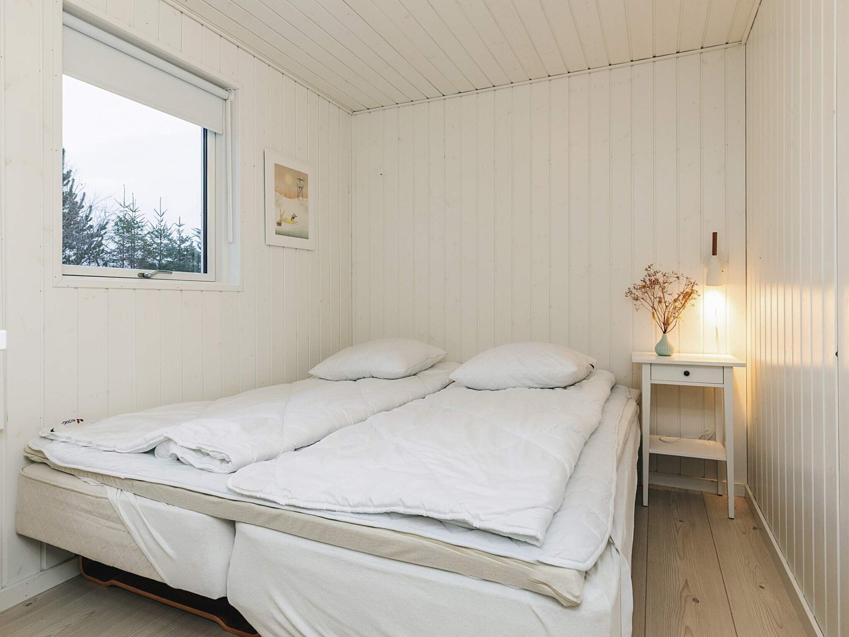 Une petite pièce aux murs recouverts de lambris blancs, comprenant un lit, une table de chevet avec une lampe et une fenêtre donnant sur des arbres.