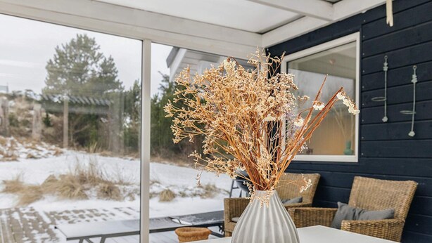 A white vase with dried flowers on a table inside a room with a large window. - 5 Star Holiday Home in Blokhus (Blokhus)