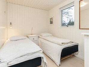 A room with two beds, a nightstand, a wall-mounted lamp, a window with a view of trees, and a framed picture on the wall. - 5 Star Holiday Home in Blokhus (Blokhus)