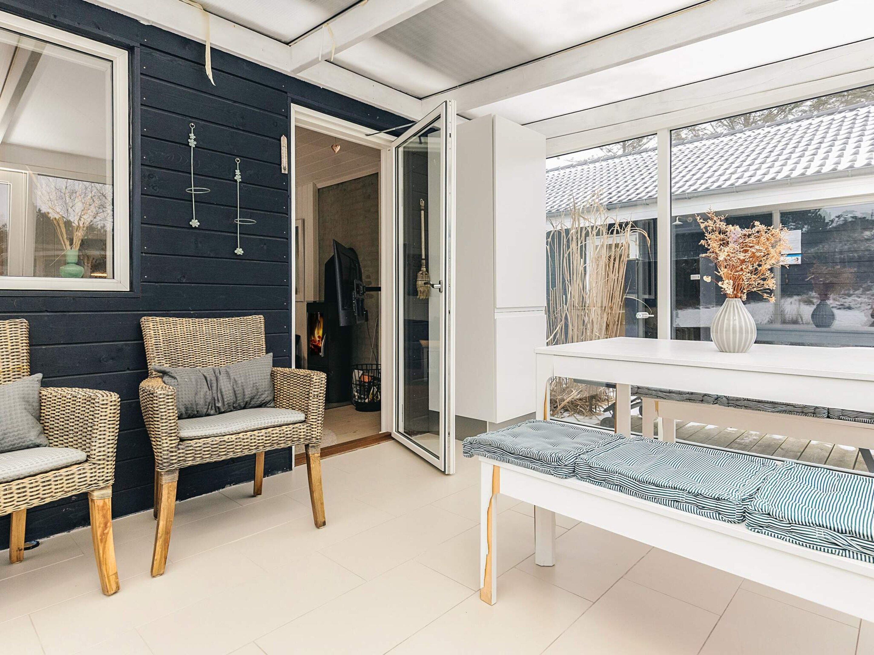 A modern interior with wicker chairs, a white bench, and a glass door leading to a room with a fireplace.