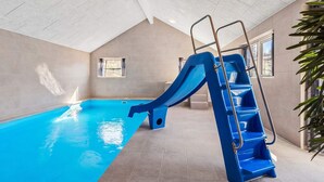 En inomhuspool med en blå vattenrutschkana och en stege, kaklade väggar och fönster.