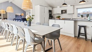 Ett modernt kök med vita skåp, en matplats med ett långt bord och stolar, hängande lampor och utsikt över utomhusmiljön genom ett takfönster.