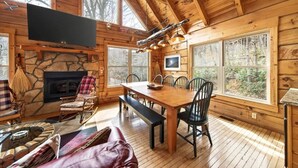 L’intérieur d’une cabane en rondins, avec une cheminée en pierre, une table à manger en bois et de grandes fenêtres.