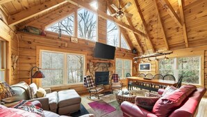 L’intérieur d  ’une cabane en rondins, avec une cheminée, un coin salon, une table à manger et de grandes fenêtres.