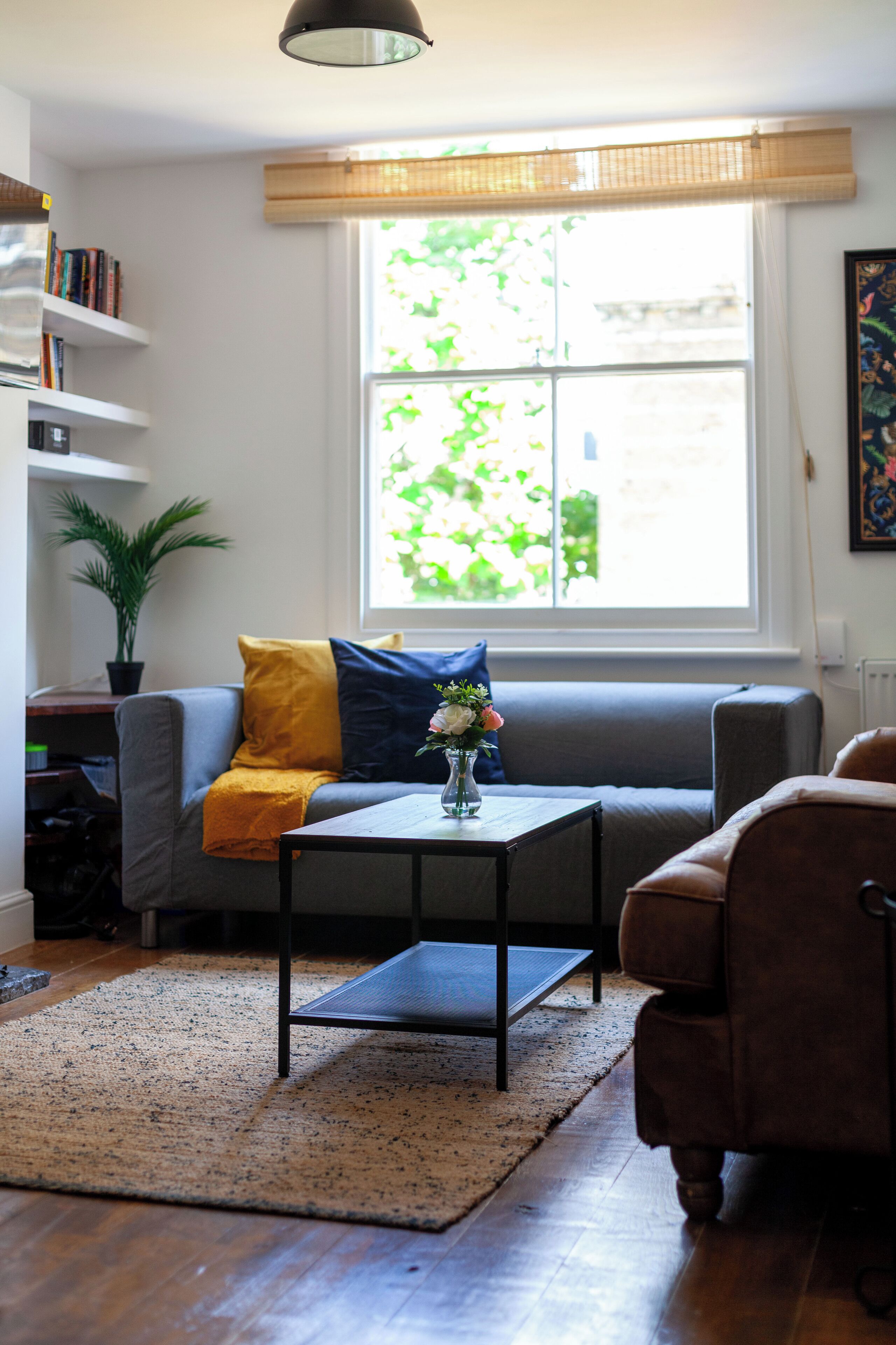 A living room with a grey sofa, a brown armchair, a small table with a vase of flowers, a bookshelf, and a large window with a view of greenery.
