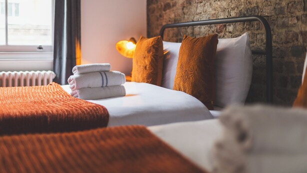 Uma cama bem arrumada com estrutura de metal, uma parede de tijolos e uma janela com luz natural.