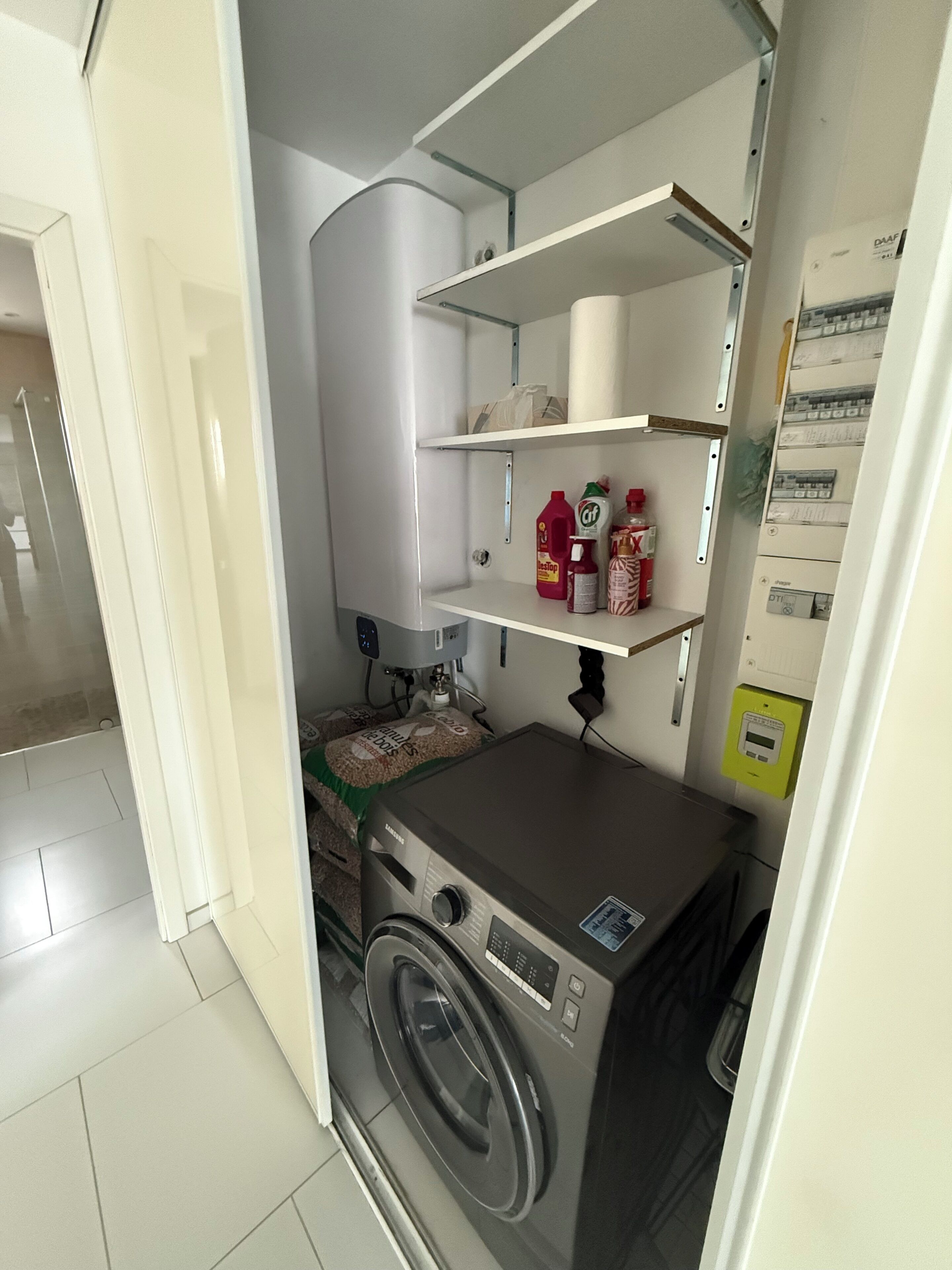 Une petite buanderie comprenant un lave-linge, des étagères avec des produits de nettoyage et un chauffe-eau.