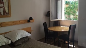 Una camera da letto con un letto, una testata in legno, un tavolo rotondo in legno con sedie, una finestra vista sul verde e una lampada fissata al muro.