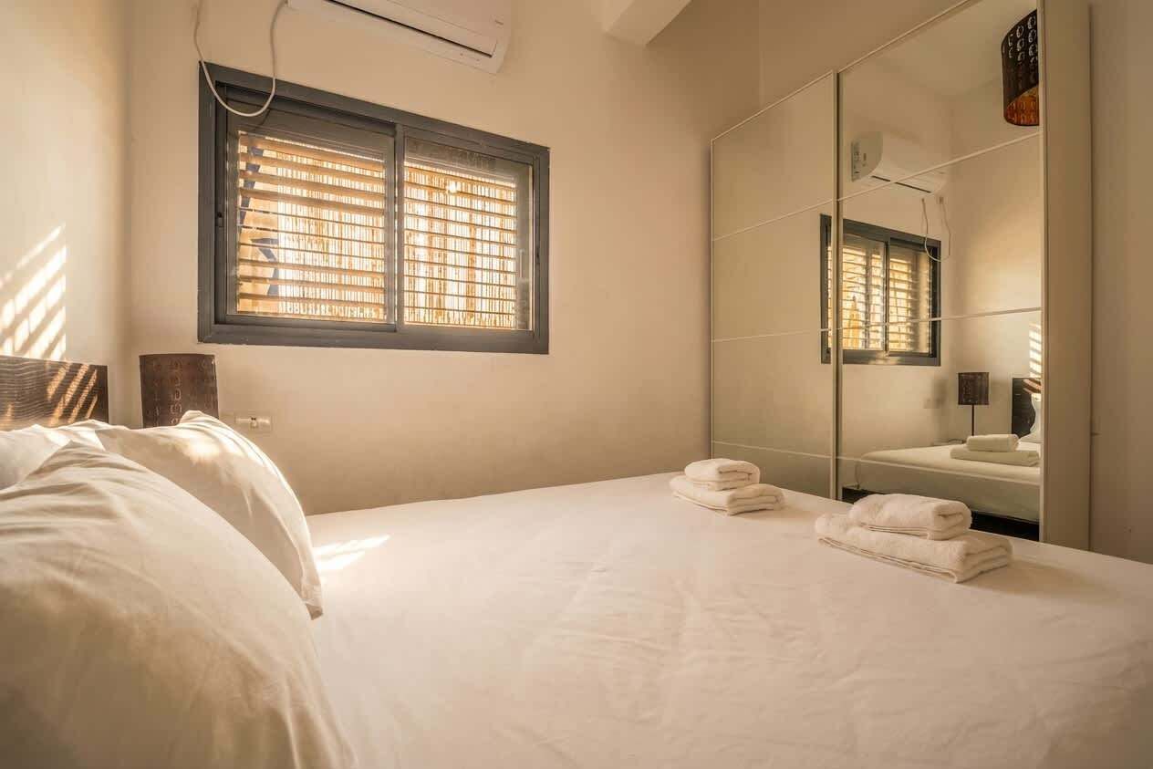 A bedroom with a bed, a large mirror, a window with blinds, and a bathroom visible in the reflection.