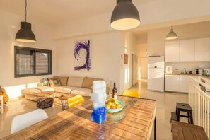 A modern living room with a wooden dining table, a sofa, and a kitchen in the background. - 3BR Garden & Cinema · Near Beach | By SweetyGuest (Tel Aviv-Yafo)