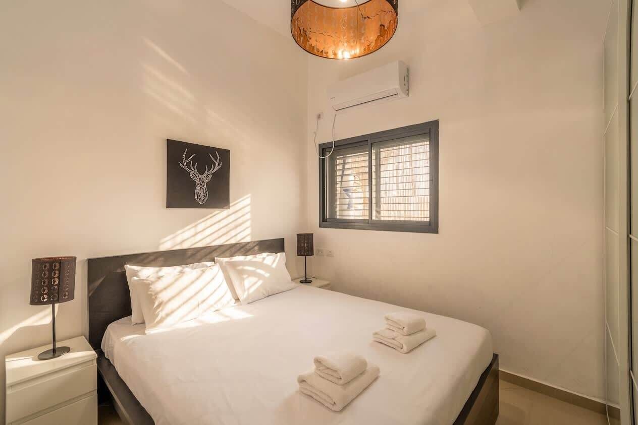 A hotel room with a bed, a nightstand, a wall-mounted air conditioner, a window with blinds, and a ceiling light fixture.