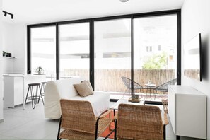 Une pièce moderne et bien éclairée, dotée d’une grande fenêtre, d’un lit, de fauteuils en osier, d’une petite table et d’un coin cuisine.