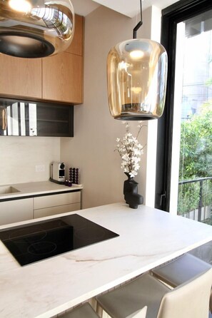 A modern kitchen with a white countertop, black induction cooktop, wooden cabinets, and a hanging pendant light. - Stylish New 2BR • Near Beachfront | By SweetyGuest (Tel Aviv-Yafo)
