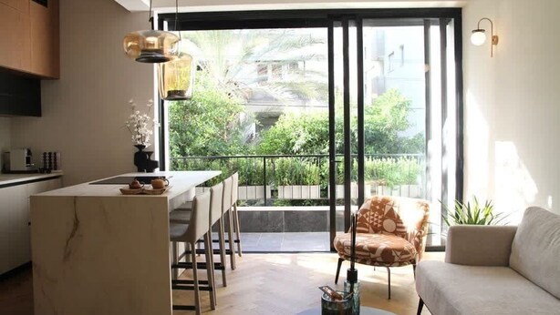 Eine moderne Küche mit einer Marmorinsel, einem Essbereich mit Stühlen, einer Couch und einer großen Schiebeglastür, die auf einen Balkon mit viel Grün führt.