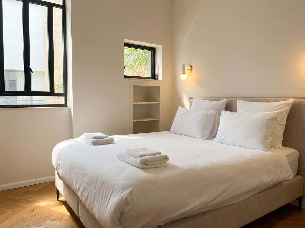 Ein ordentlich bezogenes Bett mit weißen Leinen und Handtüchern, ein kleines Fenster mit Blick auf Grünflächen und ein an der Wand befestigtes Regal.