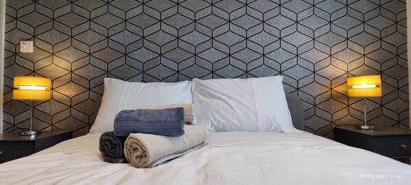 Een opgemaakt bed met witte en beige lakens, twee opgerolde handdoeken, en een hoofdbord met een geometrisch patroon.