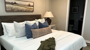 Un lit bien fait, avec une couette blanche et plusieurs oreillers, une lampe sur une table de chevet et un tableau au mur.