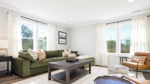 Un salon avec un canapé vert, une table basse en bois foncé et un fauteuil en cuir marron. Il y a deux fenêtres avec des rideaux blancs, et un tableau encadré accroché au mur.