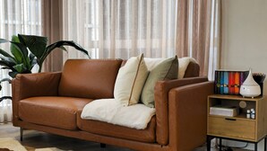 A brown leather sofa with white and green cushions, a small wooden side table with books and a white vase, and a large potted plant. - Deluxe 2BR apt Excellent Location Cityviews & Pool (Melbourne)