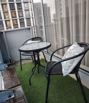 A balcony with a glass table and two chairs, artificial grass flooring, and a view of a cityscape through large windows. - Deluxe 2BR apt Excellent Location Cityviews & Pool (Melbourne)